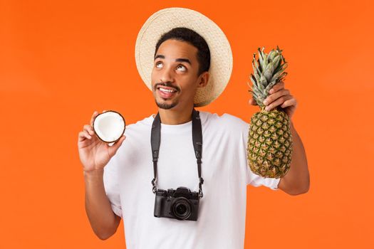 Happy dreamy handsome male tourist enjoying vacation, looking at bright sun partying on beach, holding coconut and pineapple, making cocktail, standing orange background with camera