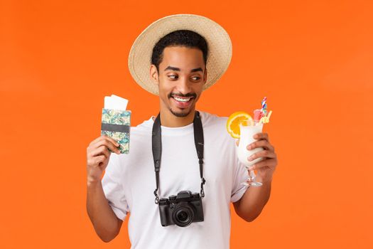 Waist-up portrait satisfied african-american male in white t-shirt and hat, looking with desire and delight cocktail, holding passport with tickets, travelling on vacation, orange background