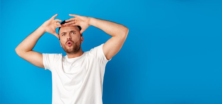 Portrait of a confused young man holding hands on his head over blue background