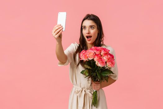 Emotions, beauty and romance concept. Attractive excited, alluring brunette female holding beautiful roses, received flowers and taking selfie with amazed surprised expression, pink background.