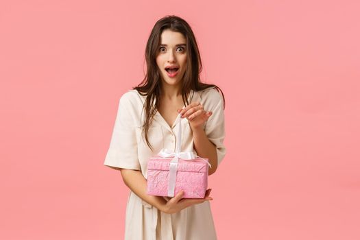 Excitement, tenderness and celebration gesture. Feminine surprised and excited alluring brunette girl in dress, wrapping gift, look amused and wondered, holding cute present box, pink background