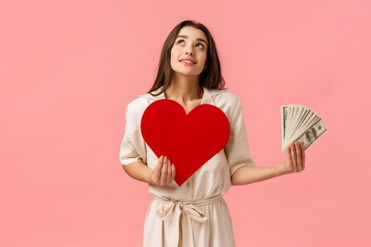 Dreamy young and cute girl imaging things, wanting find true love dont care money. Attractive alluring woman looking up thoughtful and smiling, holding heart card and cash dollars, pink background