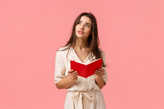 Thoughtful and inspired, creative female student having inspiration, looking up dreamy and wondered, holding red notebook, learning or preparing to class, standing pink background