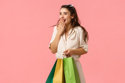 Excited and fascinated cute feminine brunette female in dress, holding shopping bags, wonder around mall, gasping excited cover mouth seeing something wonderful, pink background
