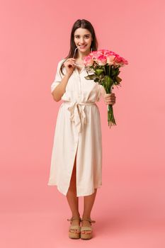 Full-length vertical portrait sensual, romantic and coquettish young woman seeing something interesting, looking with temptation or desire, holding bouquet flowers, received roses, pink background