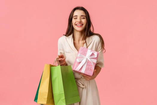 Carefree delighted female shoppaholic, girl received lots presents, holding shopping bag and cute wrapped gift, close eyes and smiling dreamy enjoying birthday party, standing pink background