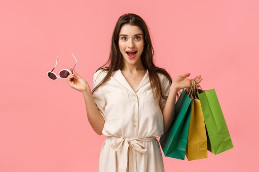 Cheerful gorgeous caucasian woman in dress, holding shopping bags and sunglasses, smiling intrigued and excited, seeing beautiful garment on stalls want buy, standing pink background