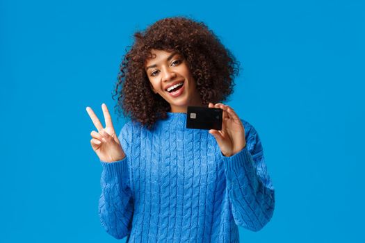 Cheerful cute african-american woman with afro haircut, tilt head show peace sign and holding credit card, got everything for holidays purchase online, standing blue background joyful