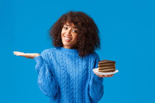 Unsure and tempting good-looking african-american girl taking care own body, looking after calories on diet, shrugging and raise hand clueless, holding plate with delicious cake, blue background