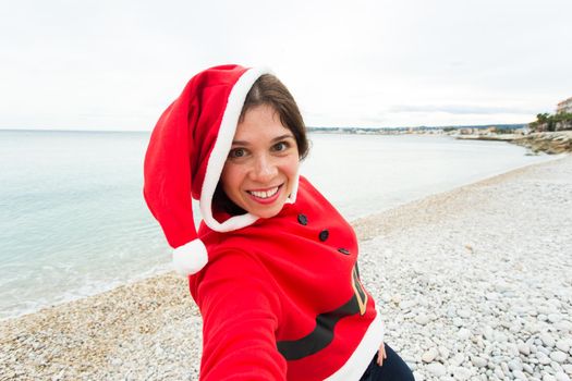 Christmas, humor and people concept - young woman in Santa suit making selfie near the sea at summer