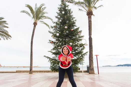 Christmas, holidays and humor concept - young happy woman have fun in christmas costume at summer near palms and christmas tree