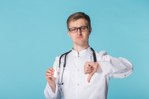 Young doctor man holding medicament showing thumb down gesture on blue background