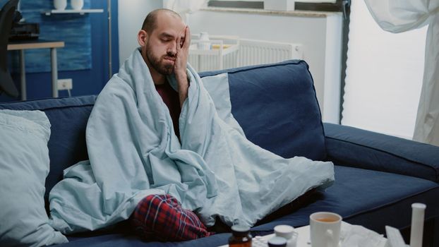 Unwell man having headache wrapped in blanket at home
