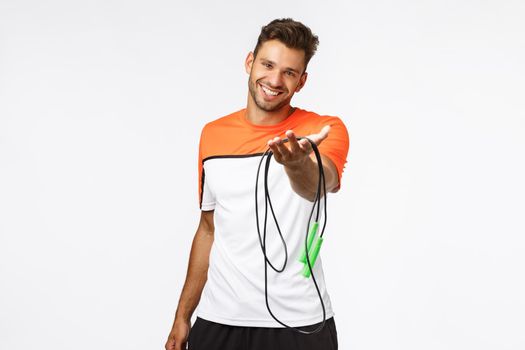 Handsome and cute smiling sportsman extend arm and suggest use his jumping rope, grinning. Charismatic fitness instructor taking care clients body, teach how workout properly, white background