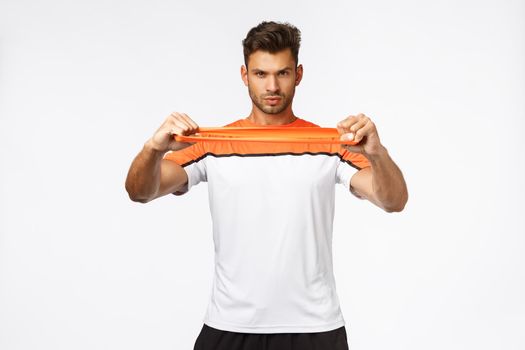 Serious, determined and motivated handsome sportsman using orange resistance band, stretch it in hands and looking focused camera, showing own strength and endurance during workout gym practice
