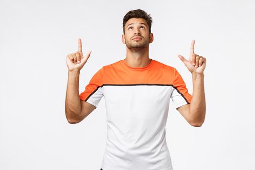 Handsome caucasian bearded sportsman, masculine guy in activewear looking up, pointing top advertisement, reading curious promo, intrigued with product advertisment, white background