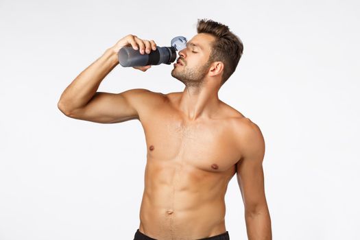 Sport, fitness and wellbeing concept. Handsome sexy young male athlete standing shirtless, holding water bottle and drinking with sassy arrogant expression, close eyes, showing naked torso, six-pack