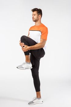Fitness, endurance and workout concept. Motivated, focused good-looking young male athlete in sportswear, raise on foot and stretch muscles to release tension, prepare body for workout exercises
