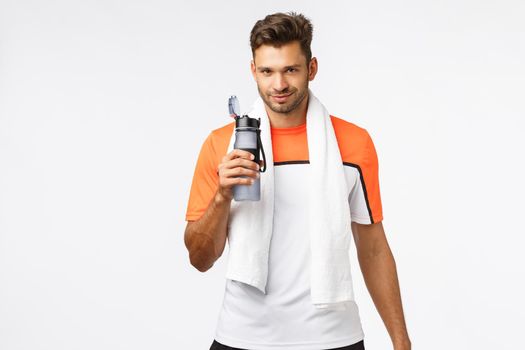 Handsome sportsman with bristle, wear activewear, towel over neck, holding bottle, drink water with satisfied smiling expression, finish good endurance practice, football player rest after match