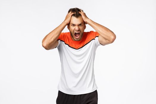 Angry, aggressive mascular sportsman looking furious, grab head in rage and fury, shouting and grimacing from anger and disappointment. Athlete lost match, team scored goal, white background