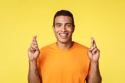 Close-up confident, hopeful, determined young man in orange t-shirt, assured he will win, cross fingers good luck, smiling happy, self-assured receive praise and positive feedback, yellow background