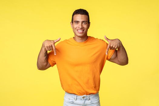 Good-looking young masculine man in orange t-shirt giving advice where buy online, pointing down smiling assertive and happy, promote advertisement, give recommendation, yellow background