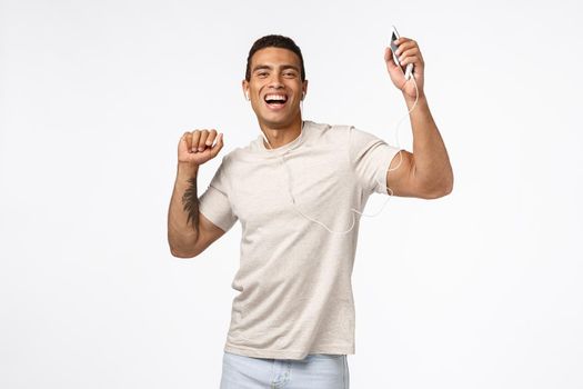 Joyful good-looking brazilian man with tattoos, raising hands up dancing carefree, enjoy awesome headphones bits, holding smartphone, wear wired earphones, smiling happy, white background