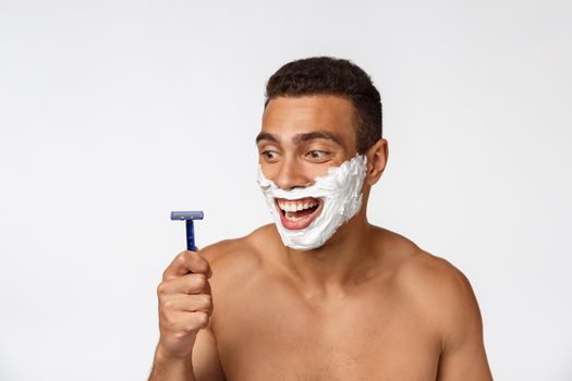 Close up of a happy naked african man with face in shaving foam holding razor isolated over gray background.