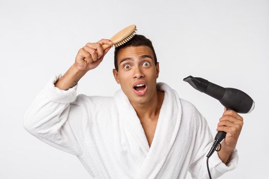 Portrait of mature funny beautiful tan-skinned african guy with curly hair in casual white bathrobe having problems with combing hair and hair dryer