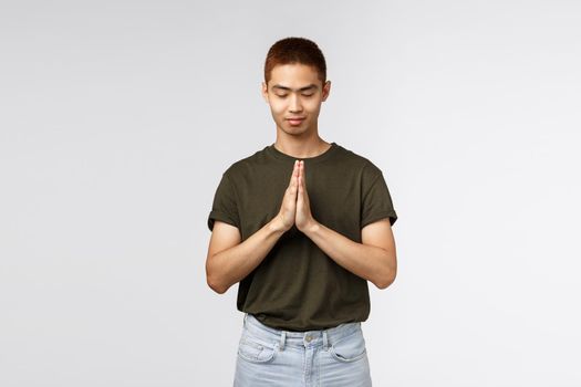 Portrait of hopeful young asian guy having faith, close eyes and smiling, clasp hands together in pray, pleading to god, want dream come true, supplicating, standing grey background