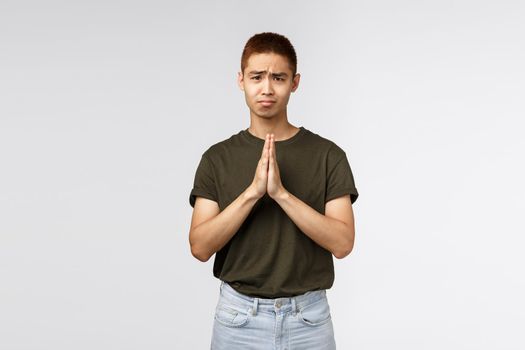 Please help me out. Portrait of hopeful clingy cute asian man asking apology, clasp hands together in pray, gloomy looking camera pleading, begging you, standing grey background