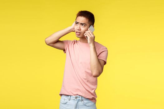 Technology, communication and lifestyle concept. Portrait of troubled, indecisive young asian guy look puzzled up, talking on mobile phone, having tough conversation, yellow background