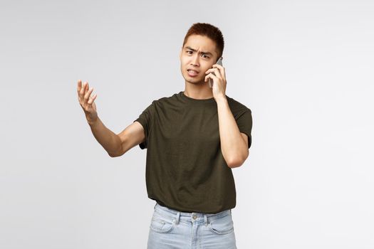 Technology, online lifestyle and communication concept. Portrait of concerned, complaining asian man shaking hands in dismay, arguing via phone call, react displeased to bad news