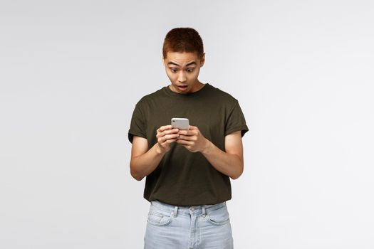 Technology, online lifestyle and communication concept. Surprised taiwanese guy, male student react to amazing news read internet social network, stare at smartphone impressed, use mobile phone