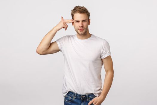 Think before act. Sassy smart good-looking handsome blond young man in white t-shirt touching temple, pointing at head or brain and smiling self-assured, telling making wise decision