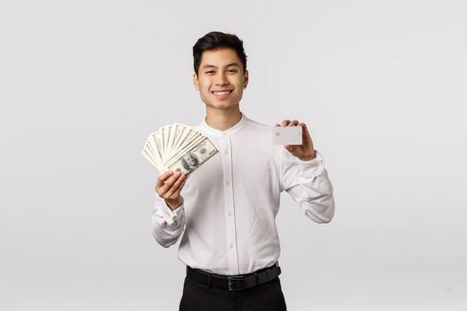 What you prefer cash or card. Satisfied good-looking asian businessman holding money and white credit card, smiling pleased, have two variants payment, being rich, standing white background