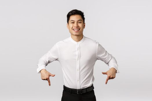 Carefree, smiling cute and joyful asian millennial guy in formal wear, shirt and pants, pointing down, inviting check out, visit event page, shopping site or corporate banner, grinning rejoicing