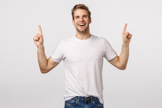 Impressed, carefree delighted, smiling young man feeling joy or happiness, grinning astounded or wondered, pointing looking dreamy up, watching amusing scene, standing white background charmed