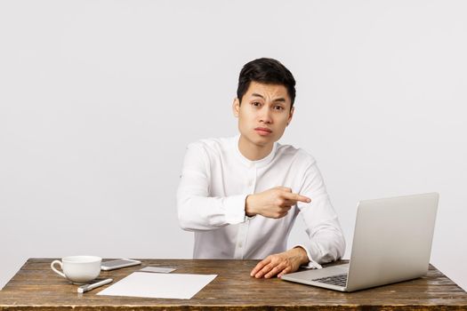 Serious displeased young asian businessman sitting office desk, pointing laptop screen and looking strict camera, frowning skeptical, scolding employee made bad report, working