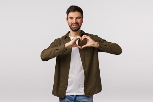 Valentines day, affection and relationship concept. Cheerful tender bearded male in coat, showing heart gesture near chest, express love or adornment, smiling cute, express gratitude or affection