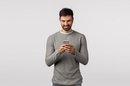 Handsome bearded caucasian guy in grey sweater making playlist, holding smartphone and listen music wireless headphones, enjoy songs using headphones, watching video in crowded place, smiling