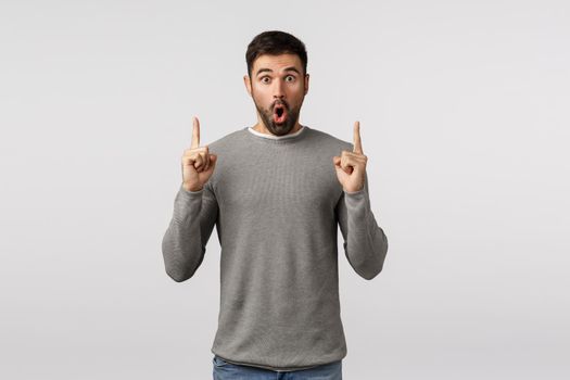 Surprised and amazed, speechless excited handsome bearded guy in grey sweater, say wow, folding lips amused, gasping astonished, pointing fingers up to turn attention, discuss impressive offer