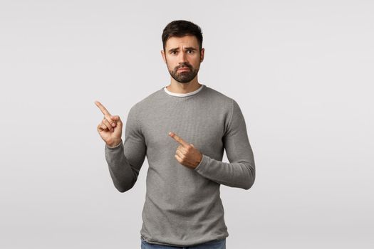 Depression, difficult situation and emotions concept. Sad, distressed and lonely bearded guy in grey sweater, frowning, look upset, pointing upper left corner with regret and disappointment