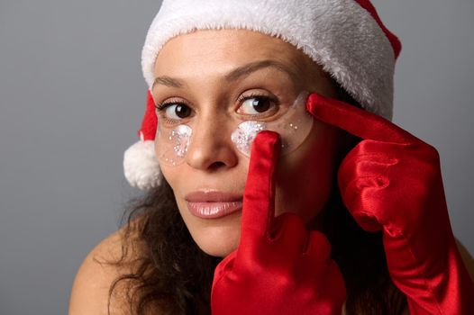 Headshot portrait of beautiful brunette woman in Santa hat wearing eye patches looking closely at camera, isolated on gray background. Skin care concept for Christmas ad of cosmetology beauty salons