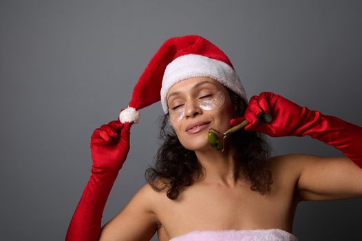 Attractive woman in Santa costume uses jade roller massager for facelift and lymphatic drainage facial massage. Skin care, cosmetology concept for christmas advertisement of beauty salons and spa