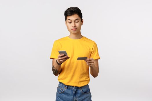Handsome asian young man in yellow t-shirt, holding smartphone and credit card, entering digit number to buy online, shopping internet, paying for application, standing white background