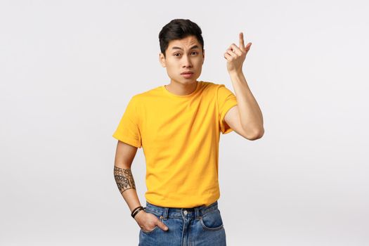 Wtf going on. Annoyed asian young man in yellow t-shirt, with tattoos, raising index finger in dismay and irritation, raising eyebrows as hearing nonsense, weird conversation, white background
