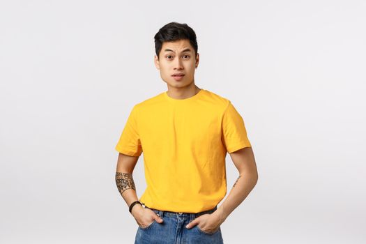 Surprised handsome asian male model didnt expact someone saying rude offensive words, raise eyebrows up in awe and wonder, hold hands in pockets, looking intense and aggressive, white background