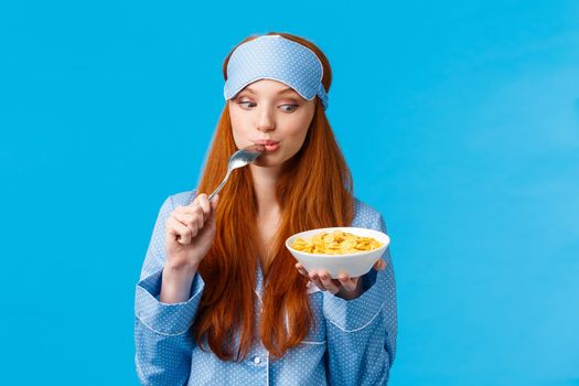 So tasty. Cute and tender feminine girl starting day with healthy diet, eating delicious cereals, licking spoon and staring plate tempting and eager eat it, standing blue background