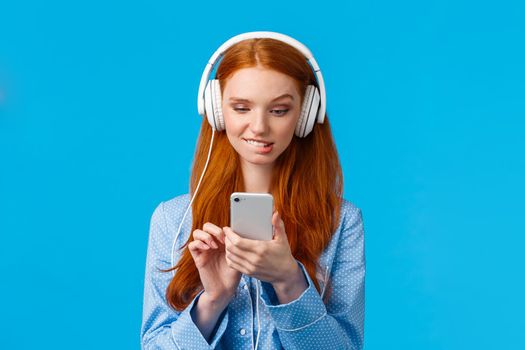 Waist-up portrait indecisive, unsure picky cute redhead female in nightwear, biting lip and looking tempting with desire smartphone screen, send risky text, listen music headphones